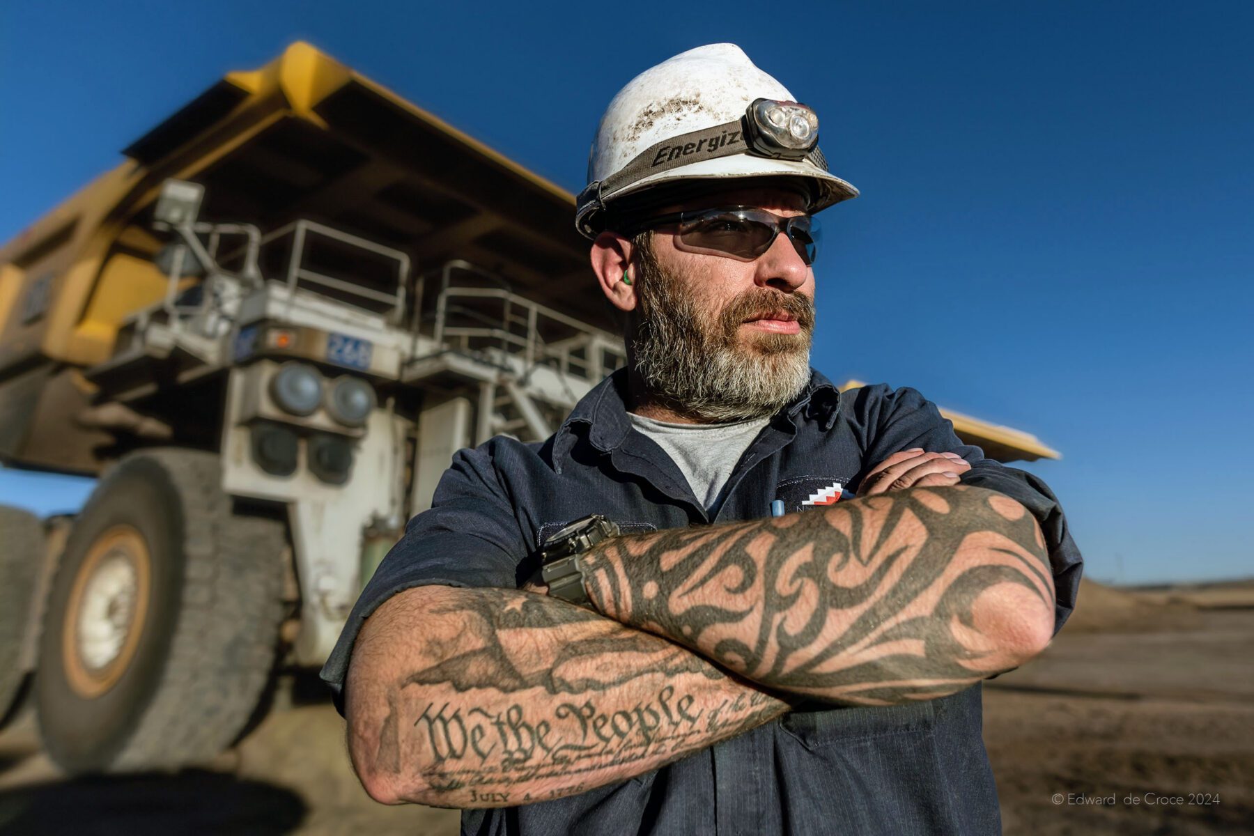 This portrait of American coal miner rendered by Denver portrait photographer, Edward DeCroce depicts a strong male driver in front of his giant truck.  Across his arm is an unmistakably patriotic tattoo with the words "We The People". The phrase, of course is the opening of the preamble to the US Constitution published in 1787. Underneath is tattooed the date 1776. 
